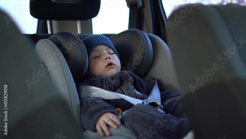 A small child sleeps in a car seat in a parking lot. Boy in a sweatshirt and hat.