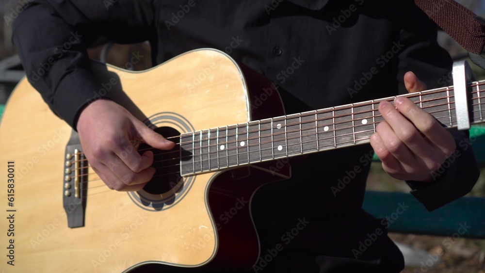 A young man plays the acoustic guitar while sitting in the park. A man plays the guitar close-up. The camera is moving.