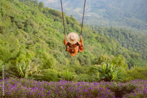 Back of beautiful womanis in brown dress is swinging on a big swing at the mountains with breathtaking view. Below is a field of purple flowers. In front is a view of green mountains.