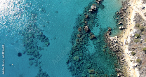 A drone flying over an empty rocky shore in clear summer weather and calm sea with clear water of different shades of blue and glare from the sun on the water. Top view.