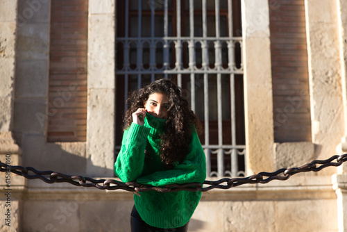 Fotografija young beautiful brunette woman with curly hair and green woollen coat is leaning on the chains surrounding the cathedral of seville in spain