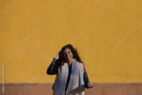 pretty young woman with curly brunette hair against a yellow background is dressed in winter clothes and wearing a scarf to protect herself from the cold. The woman is happy and having fun.
