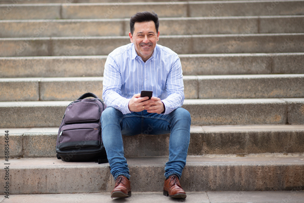 Happy mature latin man sitting outdoors on steps with bag and mobile phone