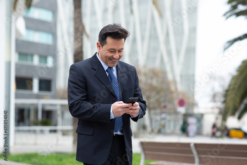 Smiling latin businessman walking in the city and using mobile phone