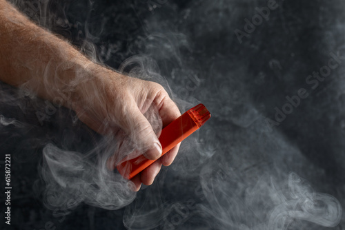 disposable electronic cigarette in a man's hand on a dark background with smoke around. The concept of modern alternative smoking, vaping and nicotine