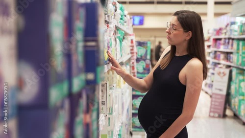 Young Pregnant Caucasian woman buying baby diapers at grocery store. Expecting female choosing diapers from shelves in the store.