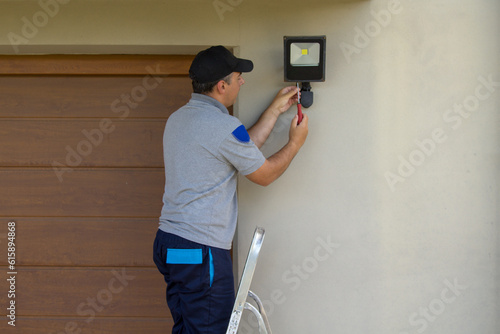 Image of an electrician on a scissor ladder using his tools to install a spotlight on a house. Illumination of the darkest parts of the house.
