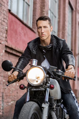 Handsome rider biker man in black leather jacket, jeans, boots and gloves sit on classic style cafe racer motorcycle. Bike custom made in vintage garage. Brutal fun urban lifestyle. Outdoor portrait.