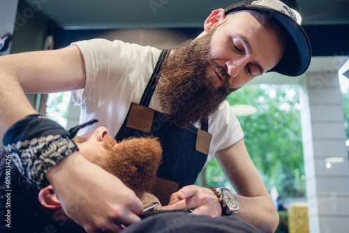 Side view headshot of a redhead bearded young man smiling, ready for shaving in the hair salon of a skilled barber with a classic straight razor in his hand