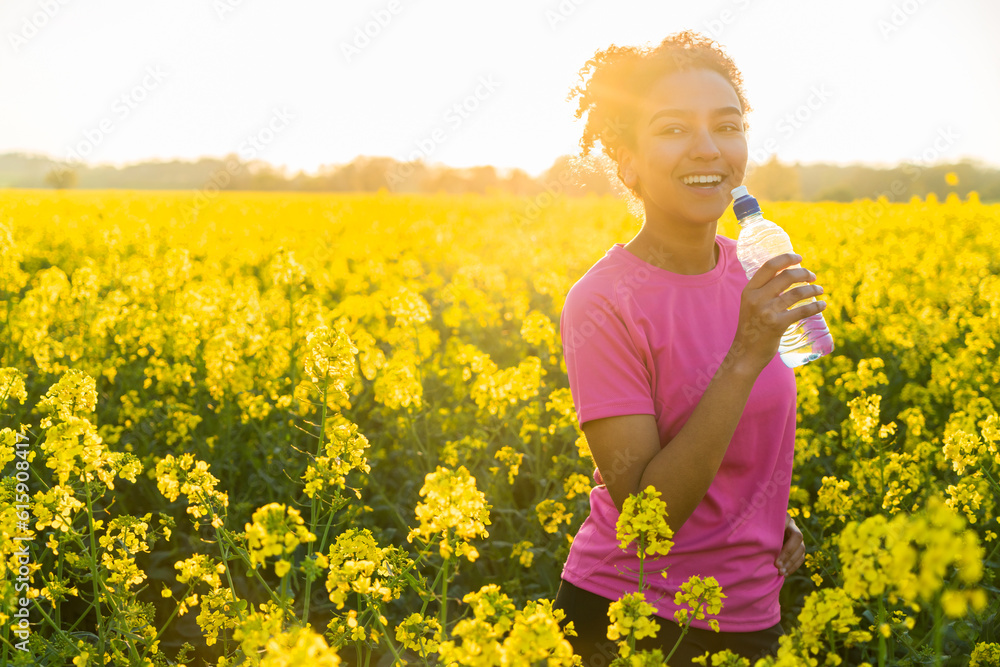 Fototapeta premium Outdoor portrait of beautiful happy mixed race African American girl teenager female young woman athlete runner drinking water from a bottle in a field of yellow flowers at sunset in golden evening su