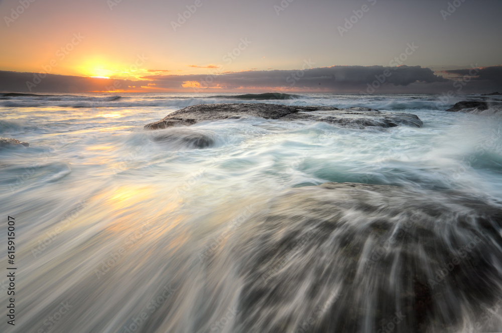 Ocean flowing rapidly over exposed rocks as the sun's glow peeks from behind clouds on the horizon.  Location, Bungan Beach, nortnern beaches Sydney Australia