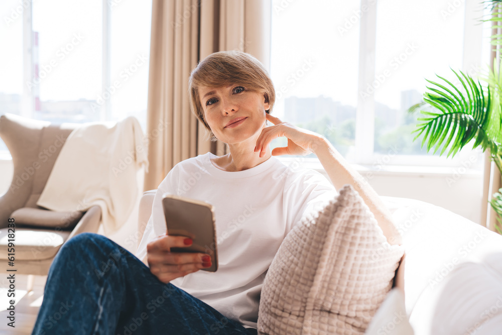 Middle aged woman with smartphone sitting on sofa in living room Middle aged woman with smartphone sitting on sofa in living room