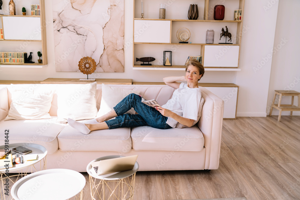 Smiling woman sitting on sofa reading magazine in apartment Smiling woman sitting on sofa reading magazine in apartment