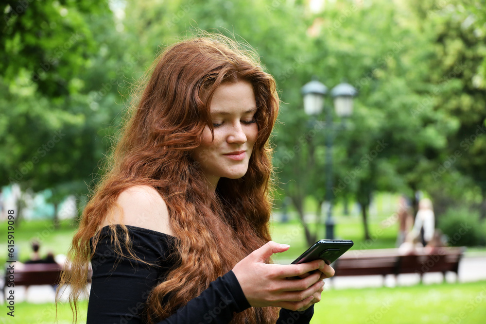 Smiling girl with long red hair and freckles using mobile phone standing in summer park. Concept of flirting and online dating Smiling girl with long red hair and freckles using mobile phone standing in summer park. Concept of flirting and online dating