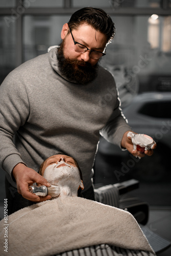 Stylish brubette barber with beard applying shaving foam onto client's face with brush in his saloon photo