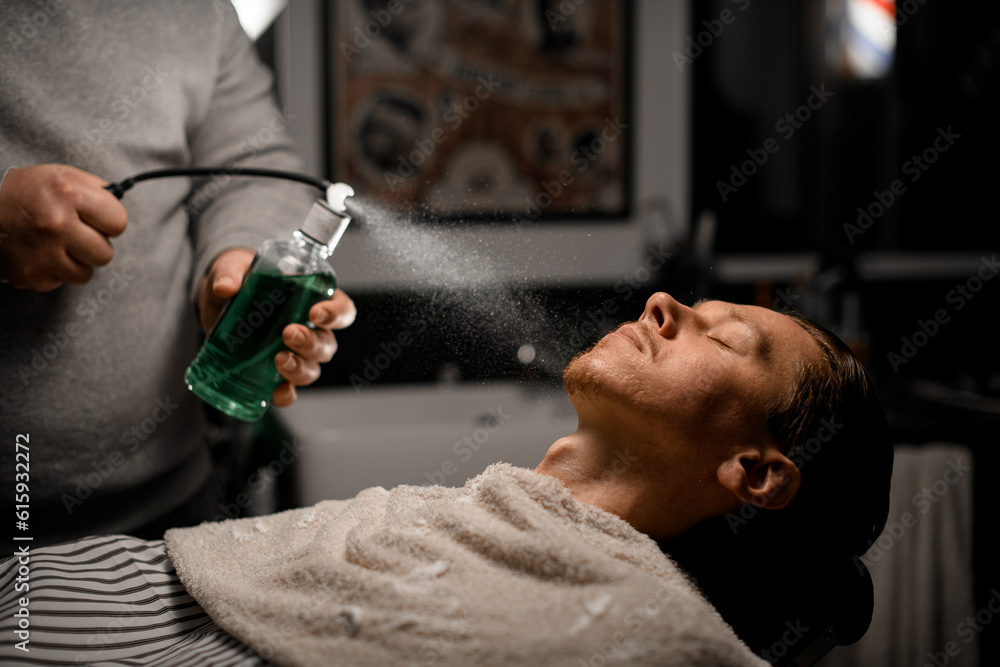 Man hairdresser sprinkles perfume on client's face in modern barbershop on black background.