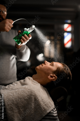 Professional hairdresser sprinkles green liquid from vintage desidned stylish bottle above client's face in modern barbershop