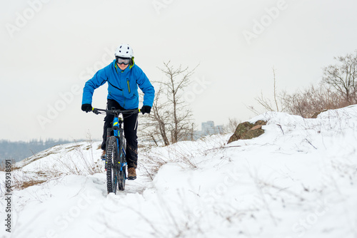 Cyclist in Blue Riding the Mountain Bike on the Rocky Winter Hill Covered with Snow. Extreme Sport and Enduro Biking Concept.