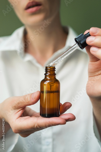 Obraz na płótnie essential oil in a brown bottle with a pipette in the hands of a woman on a green background. Women's hands with a bottle of oil and a pipette - the sphere of beauty w salonie