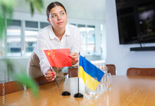 Smiling young woman preparing meeting room for international negotiations, placing national flags of China and Ukraine on table. Concept of strategic partnership and cooperation between countries