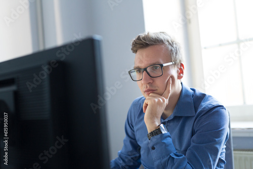 Thoughtful casual businessman wearing eyeglasses, working in office, sitting at desk, typing on keyboard, looking at computer screen.
