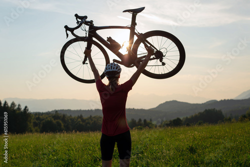 Female road cyclist raising bicycle with both arms above her head at sunset with beautiful green nature. Winning, success and achievement concept.