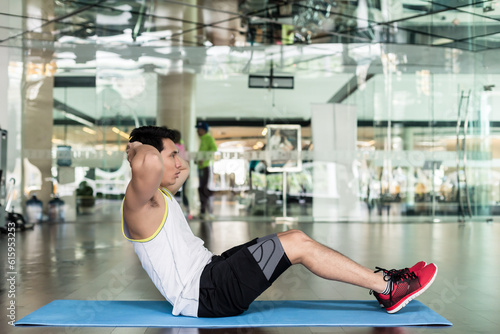 Full length side view of a fit young man sitting down on exercise mat, while doing full range crunches for the abdominal muscles at the gym
