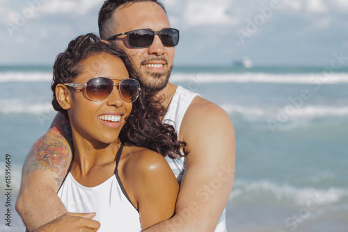 Happy handsome man embracing beautiful African-American girlfriend standing on background of ocean and looking away.