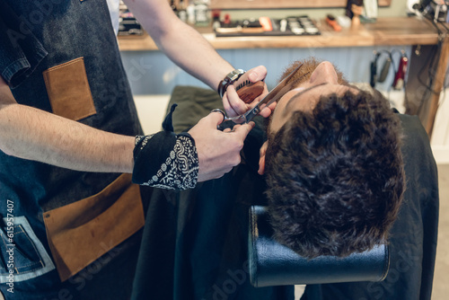 Close-up of the hand of a skilled barber using scissors, while trimming the beard of a young redhead customer in a trendy hair salon for men