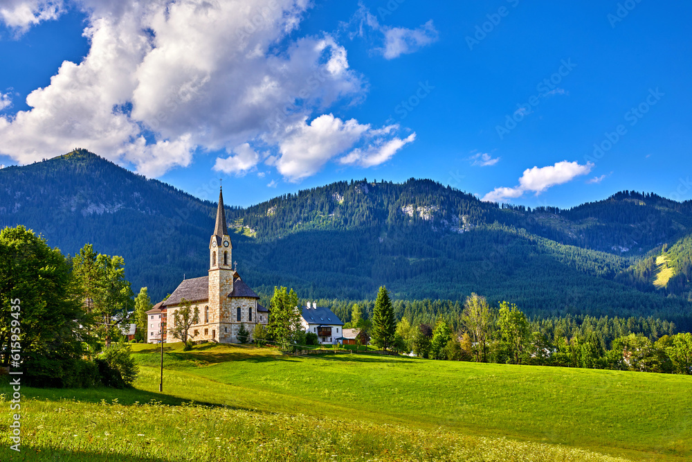 Fototapeta premium Austria. Traditional church with chapel in village among picturesque landscapes in austrian Alps mountains. Summer green lawns and fields. Knolls covered with forests and trees. Blue sky with clouds.