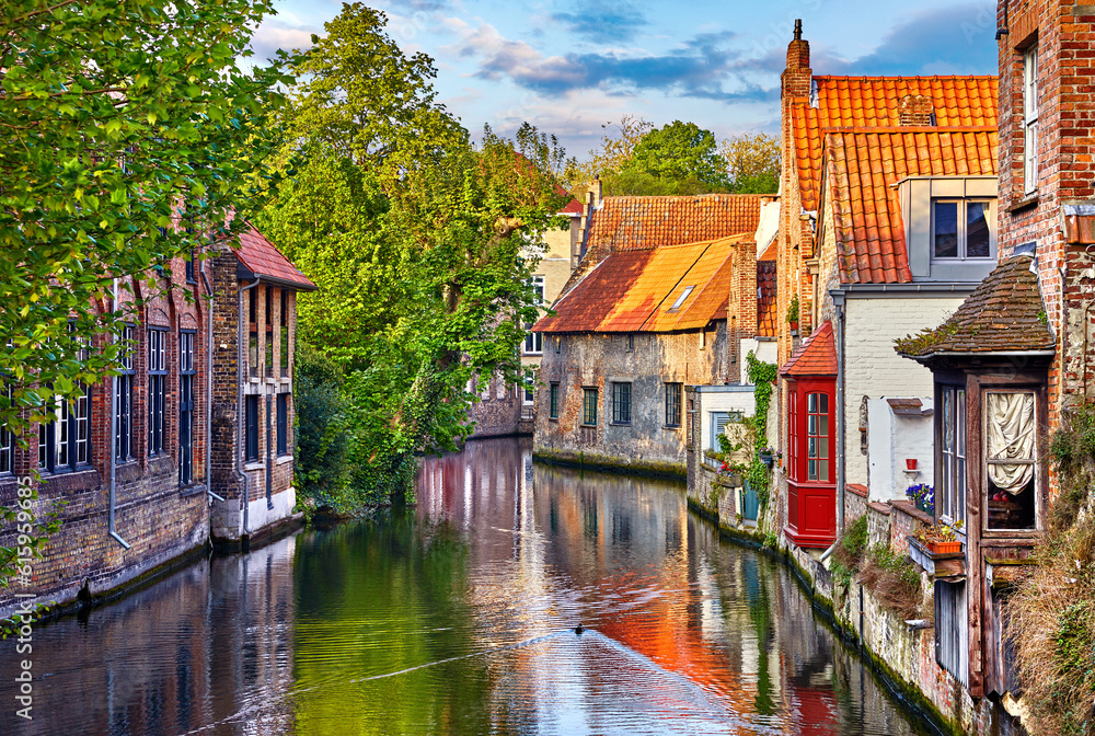 Naklejka premium Bruges, Belgium. Medieval ancient houses made of old bricks at water channel with boats in old town. Summer sunset with sunshine and green trees. Picturesque landscape.