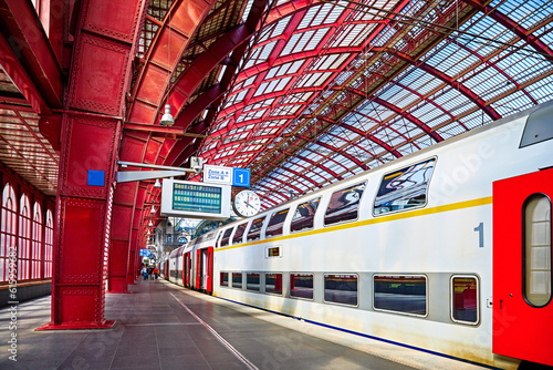Antwerp, Belgium. Central indoor railway station. Platform made of red metal constructions with clock and panel with departure or arrival schedule. Modern double decker high-speed train