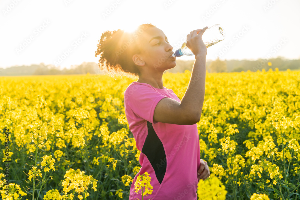 Fototapeta premium Outdoor portrait of beautiful happy mixed race African American girl teenager female young woman athlete runner drinking water from a bottle in a field of yellow flowers at sunset in golden evening su