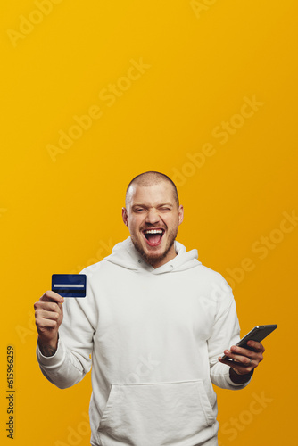 Vertical photo of excited man holding phone and credit card while shouting, standing isolated over yellow background. Empty space, mobile payment.