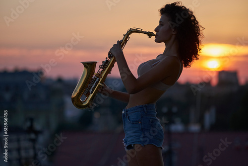 Canvas Print Young curly girl with sax on the roof at sunset