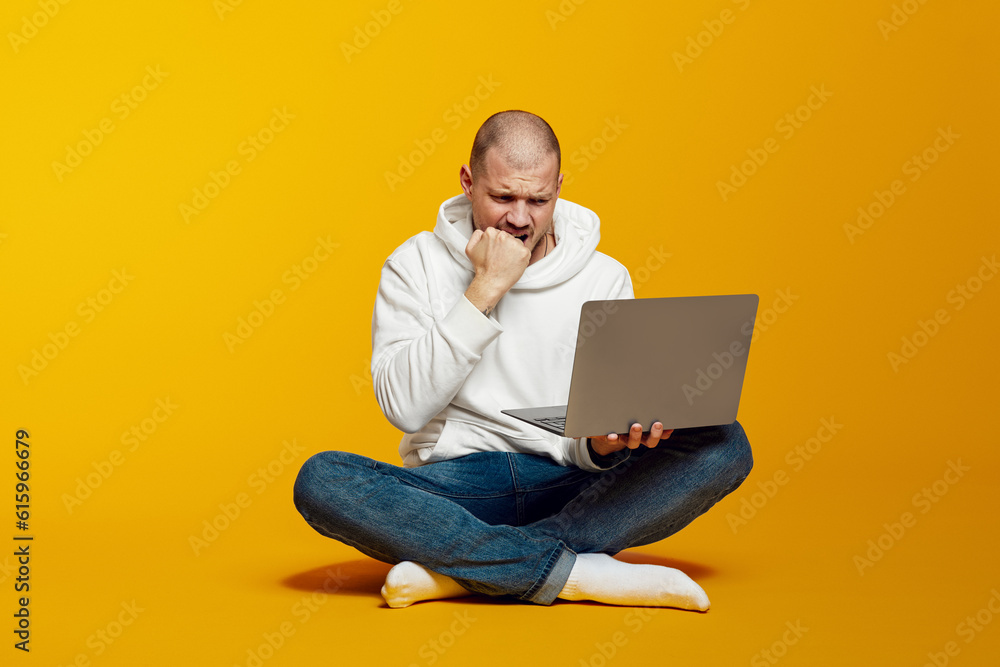 Young hipster man sitting on floor and playing in online casino and hoping his bet will win. Guy staring at laptop screen and biting his fist. Young hipster man sitting on floor and playing in online casino and hoping his bet will win. Guy staring at laptop screen and biting his fist.
