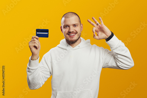 Excited young caucasian man in white hoodie holding credit bank card while showing OK gesture, isolated over yellow background