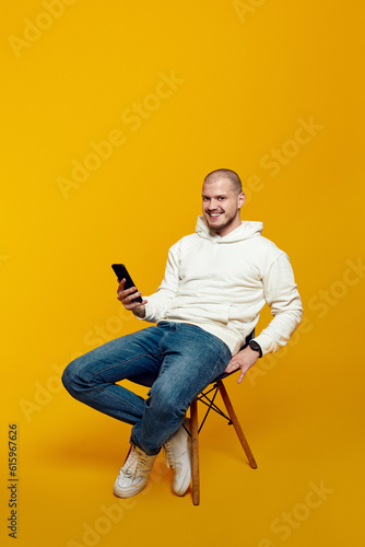 Smiling young man using smartphone while sitting on chair isolated on yellow background. Excited casual guy chatting online, browsing social media