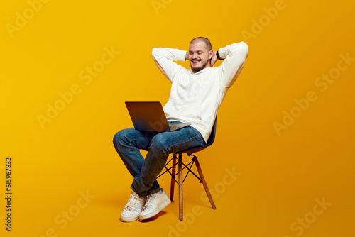 Handsome young man relaxing on chair and using laptop, happy millennial male leaning back, looking at laptop screen, isolated over yellow background