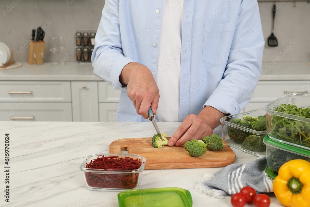 Man cutting fresh broccoli with knife near containers at white marble table in kitchen, closeup and space for text. Food storage