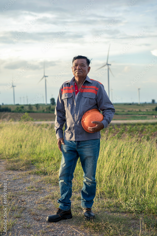 Full length portrait of senior male engineer smiling in work clothes standing in wind farm background. Elderly worker working in wind farms to generate electricity. Full length portrait of senior male engineer smiling in work clothes standing in wind farm background. Elderly worker working in wind farms to generate electricity.