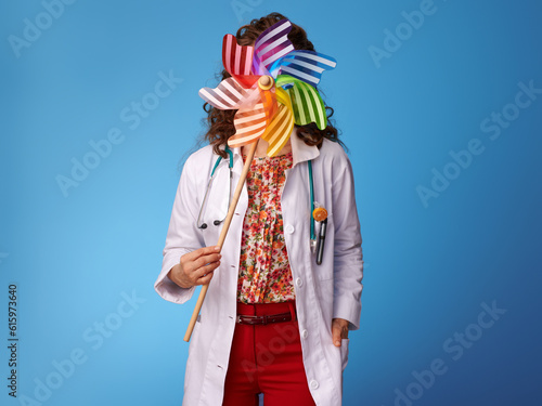 pediatrist woman in white medical robe holding colorful windmill in the front of face against blue background