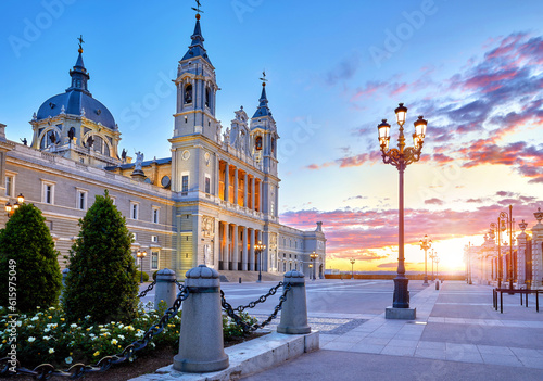 Madrid, Spain. Cathedral Santa Maria la Real de la Almudena at Plaza de la Armeria. Famous landmark with sunset sun, flowers and green bush. Street lamps with illumination and picturesque sky with clo