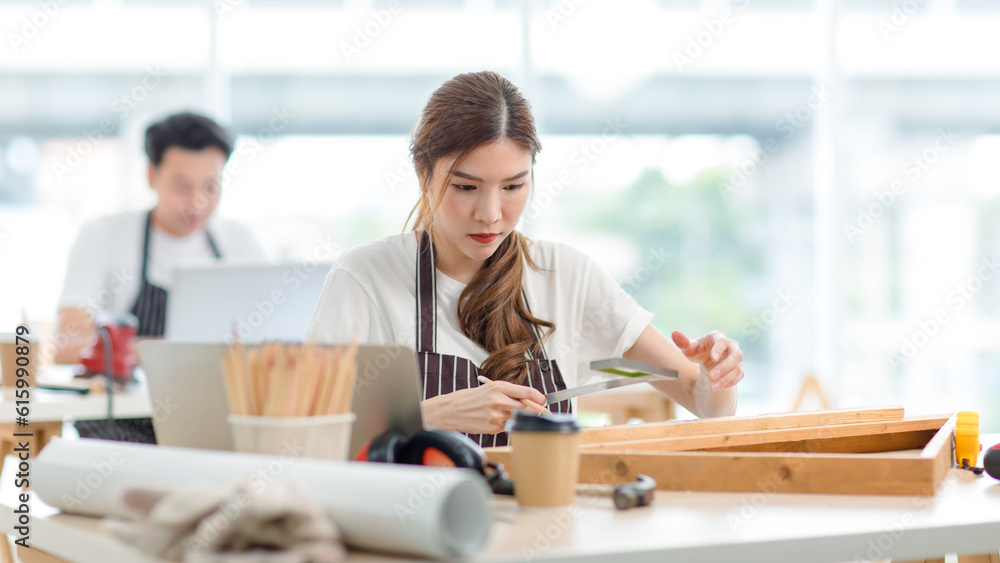 Asian professional focused female carpenter worker staff in apron sitting holding using measuring tape measure wooden sticks on workbench while male lover colleague working on blurred background