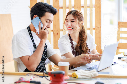 Asian professional male and female lover couple carpenter worker in apron looking pointing blueprint planning via laptop computer on wooden table with equipment in home decoration building workshop