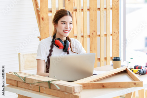 Asian professional thoughtful female carpenter worker staff with earphones in apron sitting holding pencil thinking ideas via laptop notebook computer on workbench in interior home building workshop