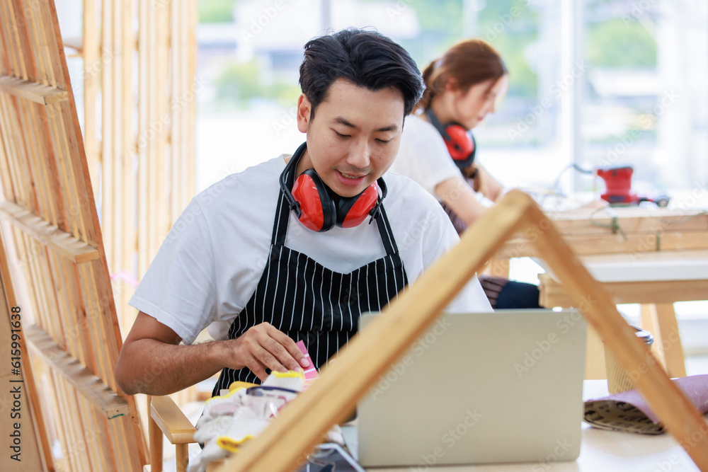 Asian professional cheerful happy male carpenter worker staff in apron with earphones sitting smiling holding video call via laptop choosing colorful color chart pattern in home building workshop Asian professional cheerful happy male carpenter worker staff in apron with earphones sitting smiling holding video call via laptop choosing colorful color chart pattern in home building workshop