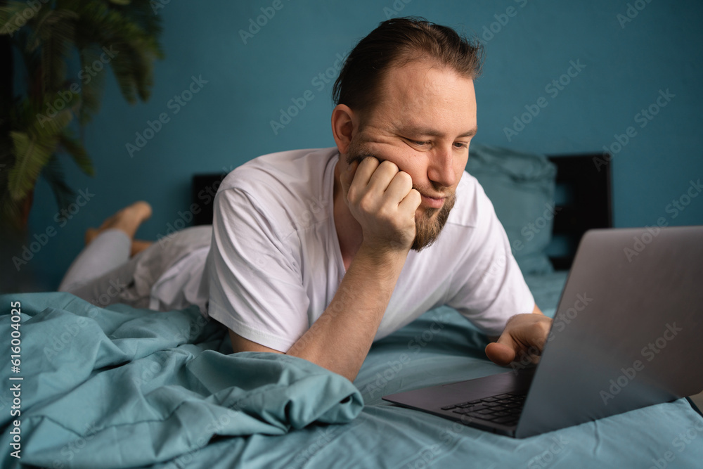 Discontented man looking at laptop lying in bed, being bored working or learning online at home. People and gadgets Discontented man looking at laptop lying in bed, being bored working or learning online at home. People and gadgets
