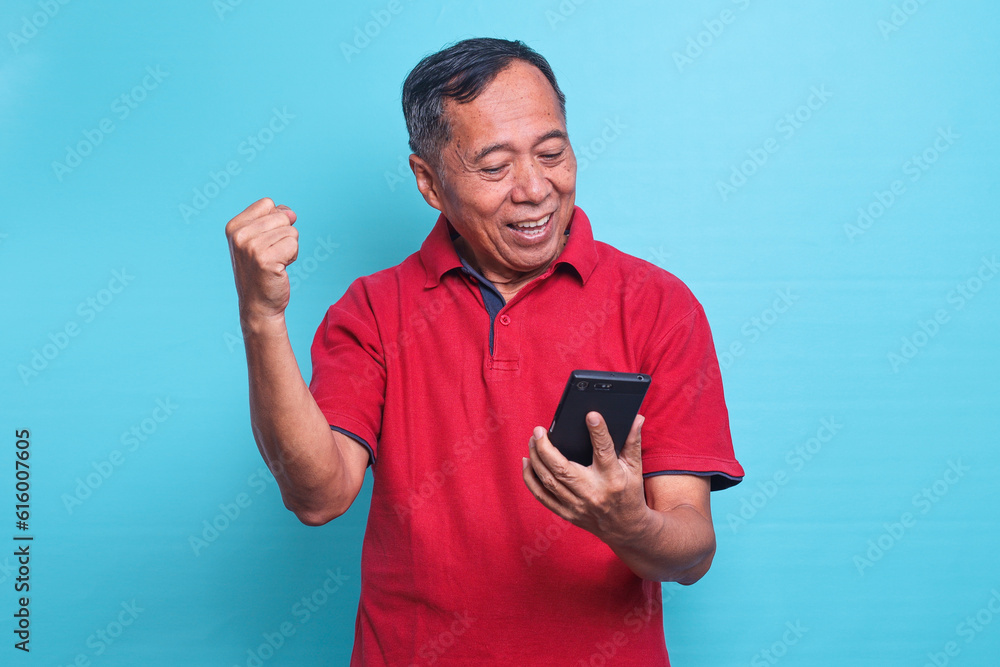 Happy excited elderly Asian man looking at smartphone gesturing winning expression getting good news over blue background Happy excited elderly Asian man looking at smartphone gesturing winning expression getting good news over blue background