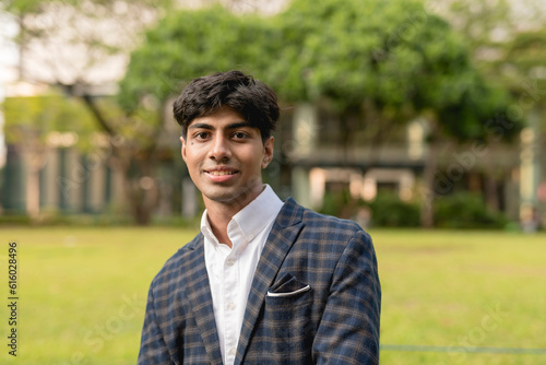 A handsome guy in a white long sleeves and blue checkered suit is smiling at the camera while standing in a greenery area. Trees, house and grass in the background.